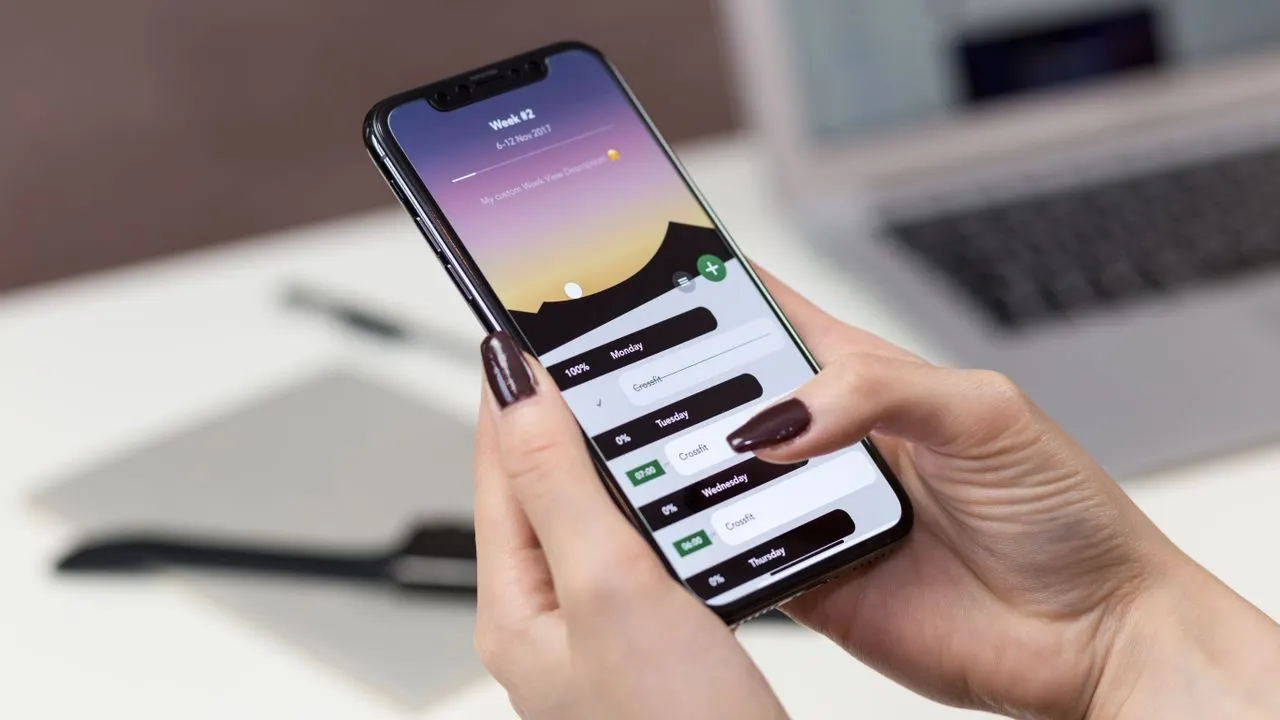 A hand holding a smartphone with apps above a laptop