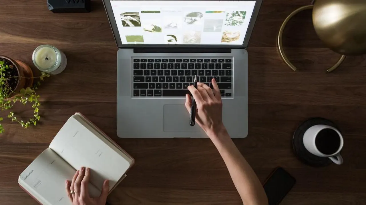 A person taking notes beside a laptop on a wooden desk