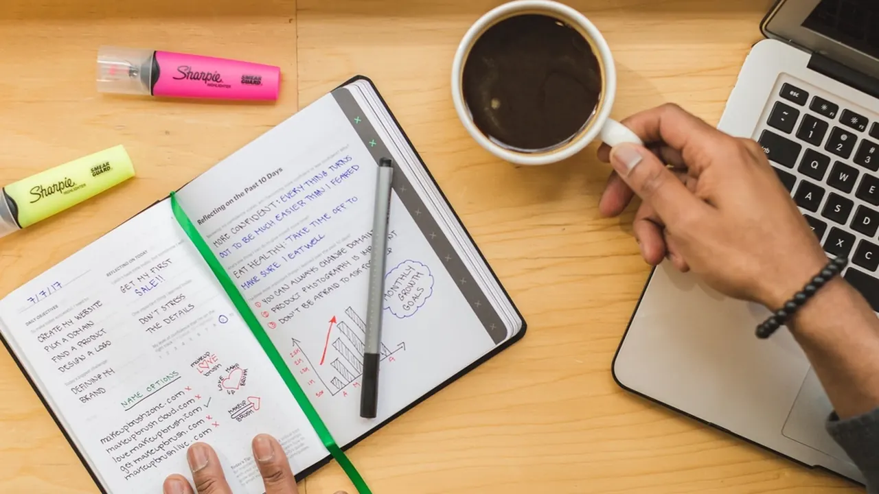 An open planner with handwritten notes beside a coffee and laptop