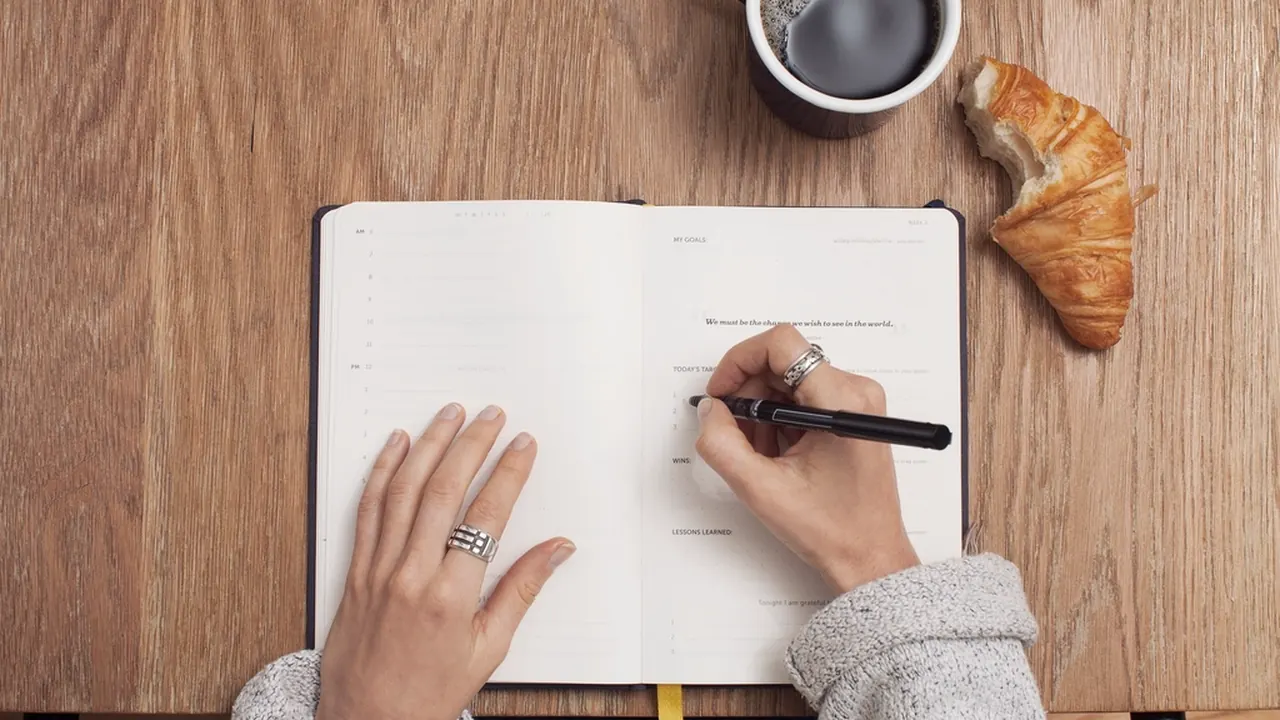 A person writing in a notebook beside coffee and a croissant