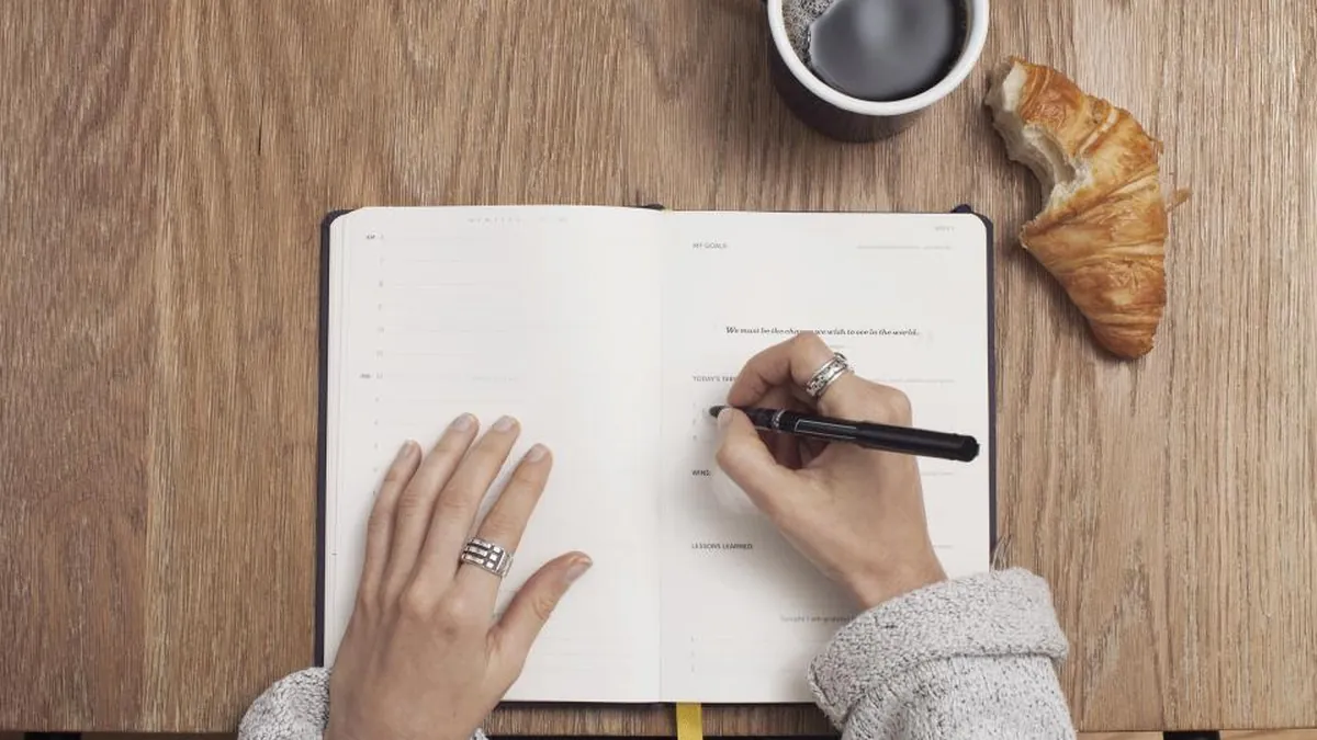 A hand writing in a journal next to a cup of coffee