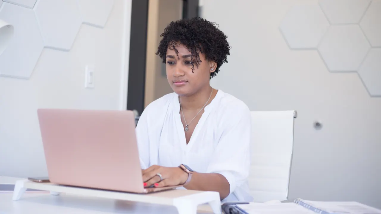 A young professional working on a laptop at a white desk in a bright office