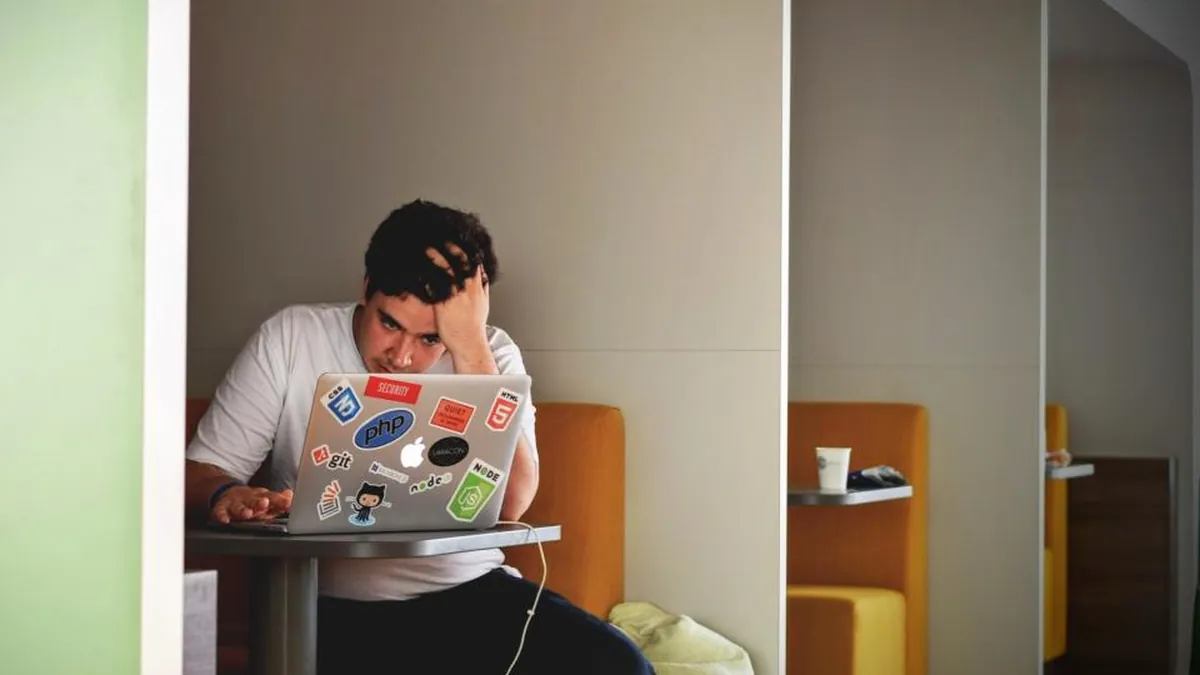 A stressed man holding his head while working on a laptop