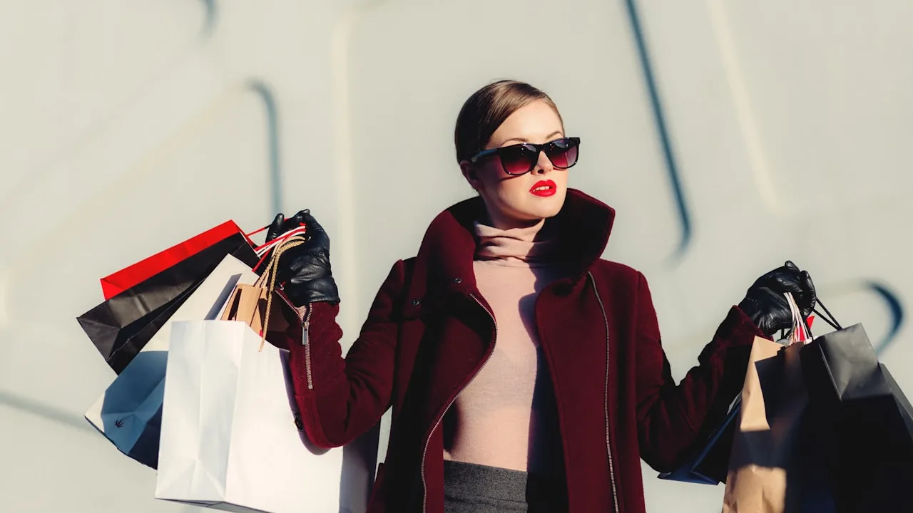 A woman in a burgundy coat carrying luxury shopping bags on a bright city street